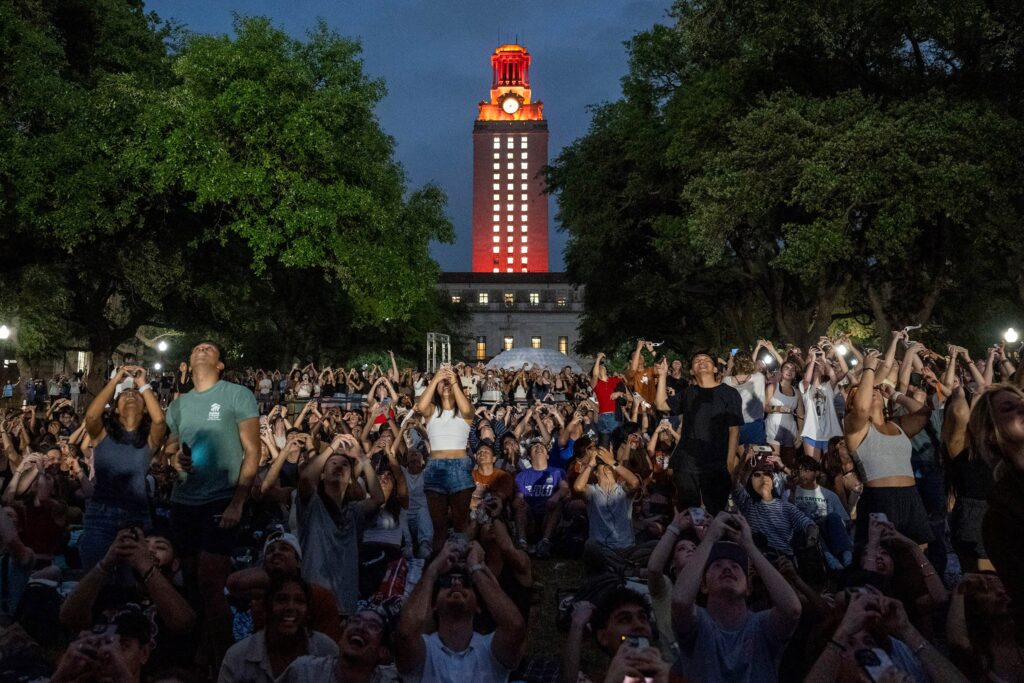 Large crowd of students, faculty and staff on campus looking up at the eclipse through longhorn eclipse glasses in front of the UT Tower glowing orange during totality.