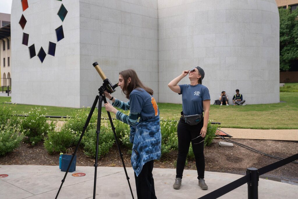 Students, faculty, staff looking up at the eclipse through glasses and one with a telescope near the Blanton Museum of Art "Austin" exhibit.
